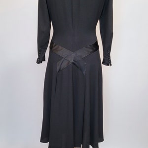 Puede incluir: Un vestido negro con un corpi&ntilde;o ajustado y una falda acampanada. El vestido tiene una faja de sat&eacute;n negro que se cruza en la espalda y se ata a la cintura.