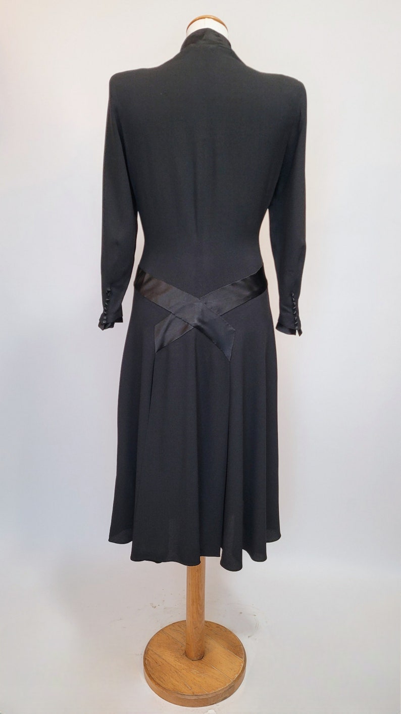 Puede incluir: Un vestido negro con un corpi&ntilde;o ajustado y una falda acampanada. El vestido tiene una faja de sat&eacute;n negro que se cruza en la espalda y se ata a la cintura.