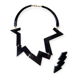 Puede incluir: Un collar geométrico negro con un diseño en zigzag. El collar está hecho de plástico y tiene un cierre dorado.