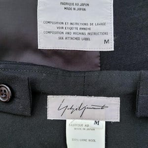 May include: A black garment featuring two white rectangular labels. The top label displays the Yohji Yamamoto signature, 'FABRIQUE AU JAPON MADE IN JAPAN', and washing instructions. The bottom label shows the signature, size M, and '100% LAINE WOOL'.
