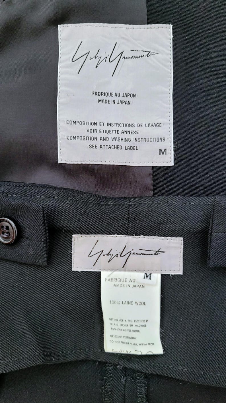 May include: A black garment featuring two white rectangular labels. The top label displays the Yohji Yamamoto signature, 'FABRIQUE AU JAPON MADE IN JAPAN', and washing instructions. The bottom label shows the signature, size M, and '100% LAINE WOOL'.