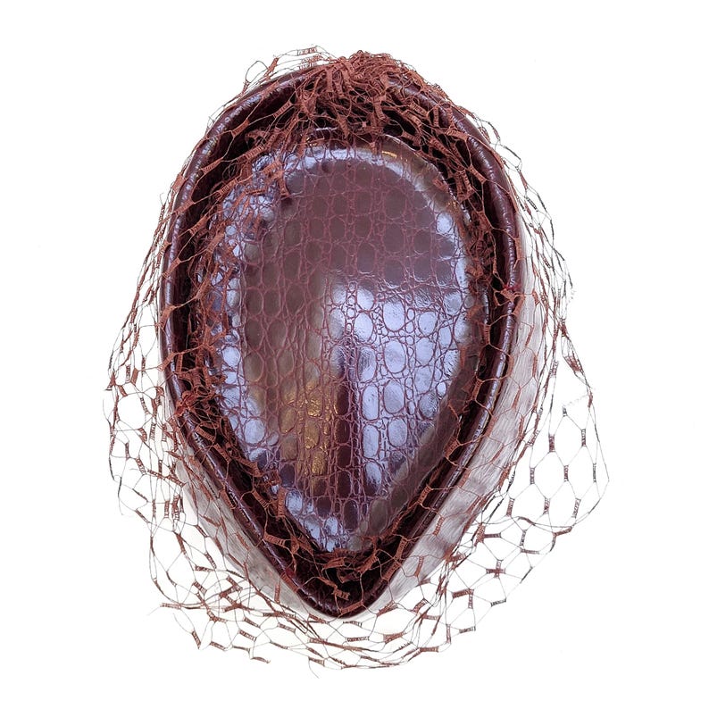 May include: A dark brown pillbox hat with a glossy, textured crocodile-skin pattern. The hat is oval-shaped and features a delicate, matching brown netting veil that drapes over the top and sides of the structured crown, creating a layered effect.