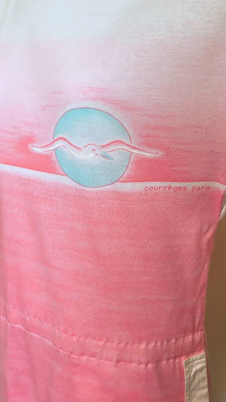 May include: A pink and white ombre t-shirt with a seagull flying over a blue circle. The text "Courr&egrave;ges Paris" is printed on the bottom right corner of the shirt.