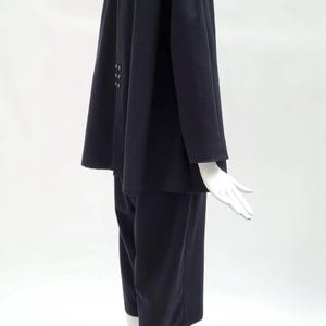 May include: A dark navy blue two-piece set consisting of a long-sleeved jacket and matching cropped trousers. The jacket features a structured collar, a flared silhouette, and a vertical row of three small buttons positioned on the back panel.