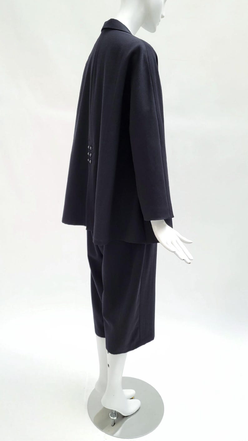 May include: A dark navy blue two-piece set consisting of a long-sleeved jacket and matching cropped trousers. The jacket features a structured collar, a flared silhouette, and a vertical row of three small buttons positioned on the back panel.