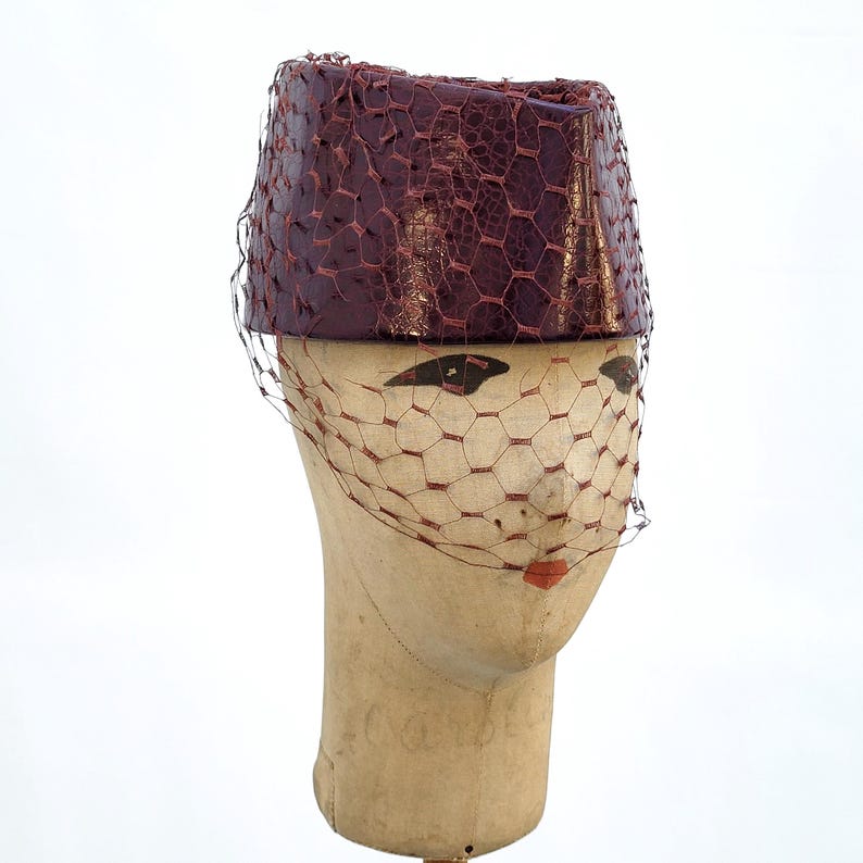 May include: A cylindrical pillbox hat in a deep burgundy colour with a textured, reptilian-like finish. The hat is adorned with a fine, matching burgundy netting veil that drapes over the front, creating a structured and layered appearance.