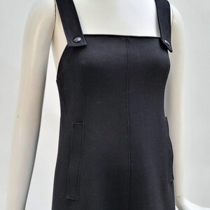 Pu&ograve; includere: Un vestito nero con scollatura quadrata e ampie spalline. L'abito presenta due tasche frontali ed &egrave; esposto su un manichino. Le spalline sono fissate con bottoni. L'abito &egrave; in tinta unita e sembra essere realizzato in un tessuto liscio.