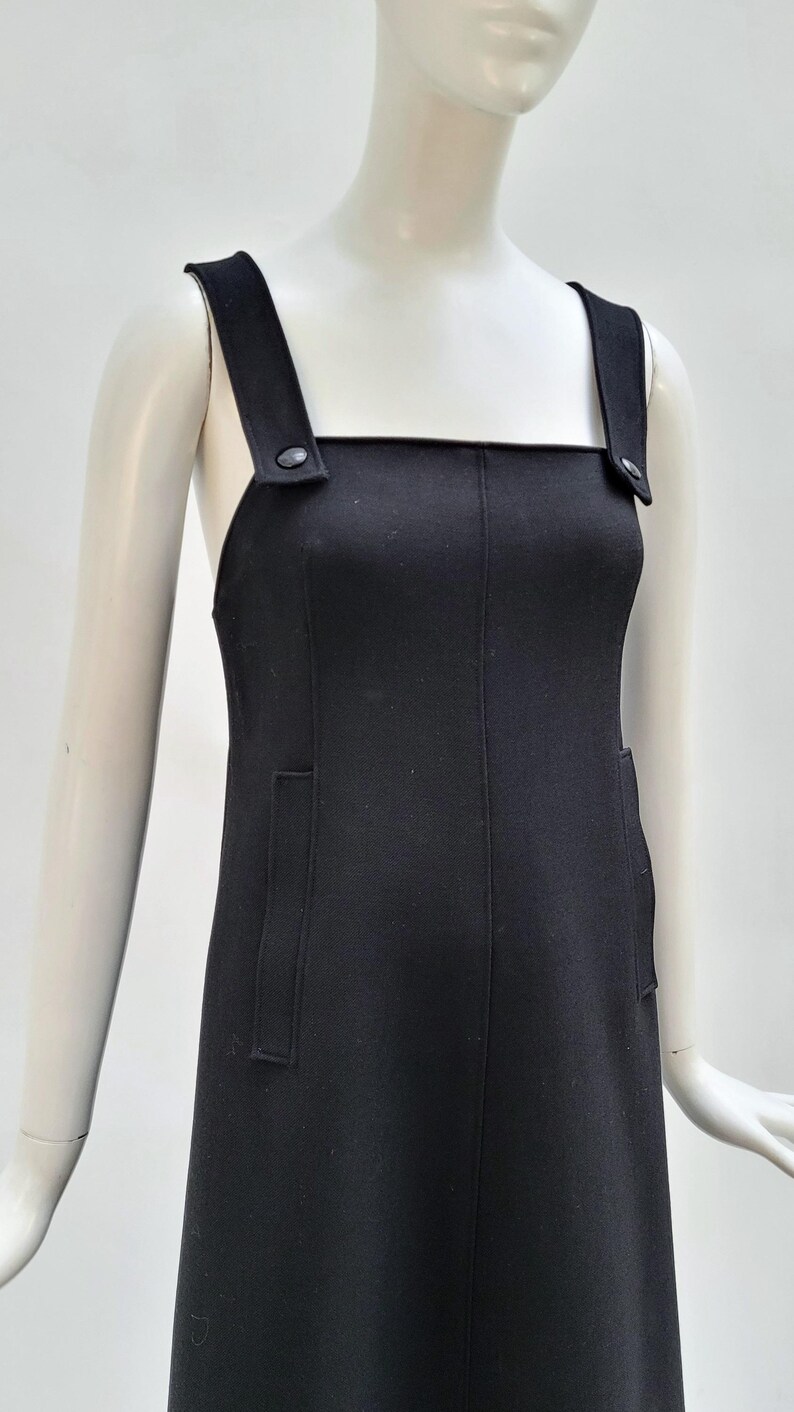 Pu&ograve; includere: Un vestito nero con scollatura quadrata e ampie spalline. L'abito presenta due tasche frontali ed &egrave; esposto su un manichino. Le spalline sono fissate con bottoni. L'abito &egrave; in tinta unita e sembra essere realizzato in un tessuto liscio.