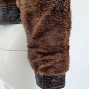 May include: Close-up of a brown fur jacket sleeve. The sleeve has a dark brown leather cuff with a button fastening. The fur is a rich, warm brown colour, with a soft, textured appearance. The jacket appears to be vintage or high-end.