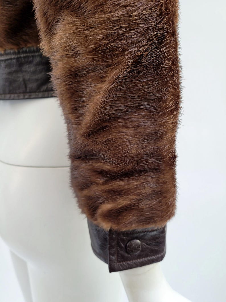 May include: Close-up of a brown fur jacket sleeve. The sleeve has a dark brown leather cuff with a button fastening. The fur is a rich, warm brown colour, with a soft, textured appearance. The jacket appears to be vintage or high-end.