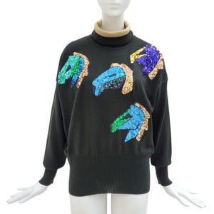 May include: Dark grey sweater with a tan turtleneck and black cuffs. The sweater features sequined abstract designs in shades of blue, green, and gold. The sweater is displayed on a mannequin.
