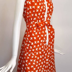 May include: A sleeveless orange dress with a white geometric pattern. The dress has a white collar, a zipper down the front, and a white belt that ties at the waist.
