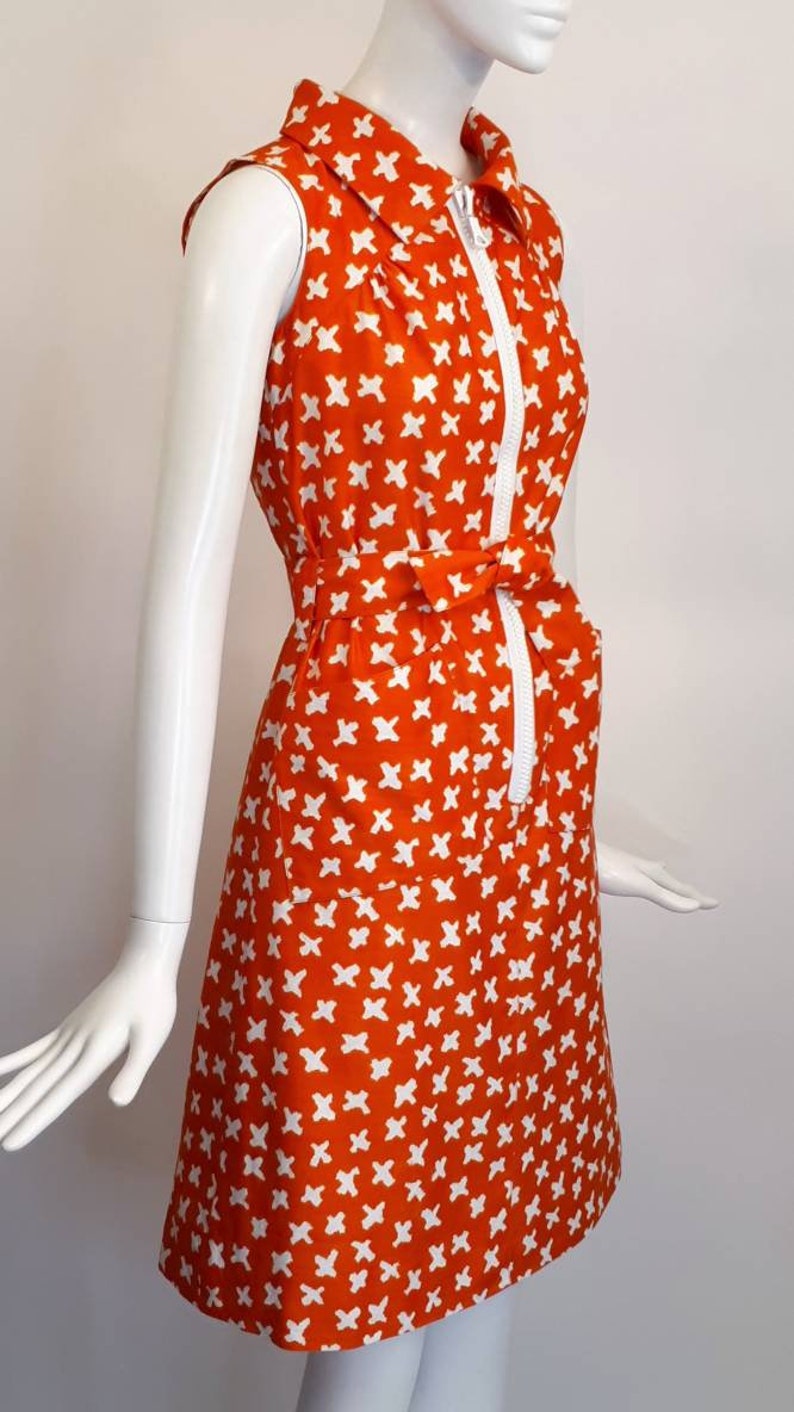May include: A sleeveless orange dress with a white geometric pattern. The dress has a white collar, a zipper down the front, and a white belt that ties at the waist.