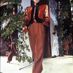 May include: A glossy, textured rust-coloured jacket with black fur trim on the collar and cuffs, paired with matching high-waisted rust-coloured trousers and a structured, shiny rust-coloured pillbox hat, creating a coordinated ensemble.