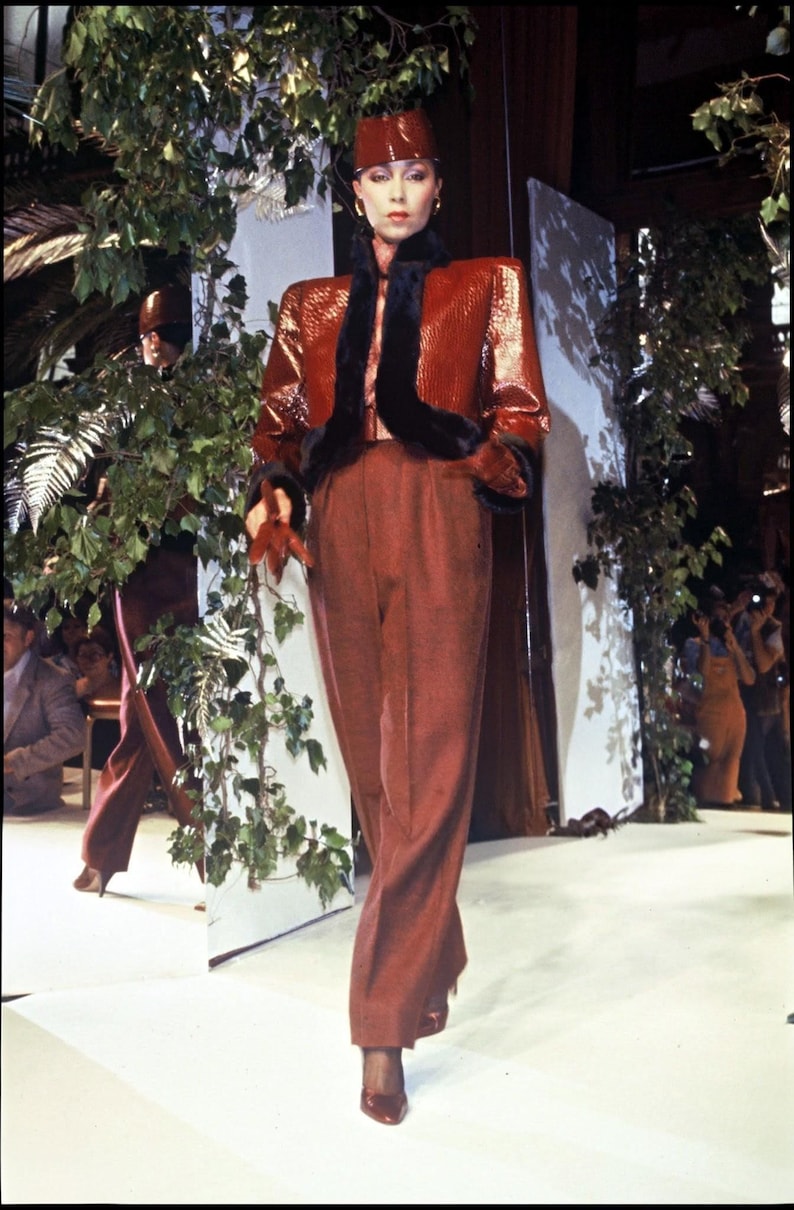 May include: A glossy, textured rust-coloured jacket with black fur trim on the collar and cuffs, paired with matching high-waisted rust-coloured trousers and a structured, shiny rust-coloured pillbox hat, creating a coordinated ensemble.