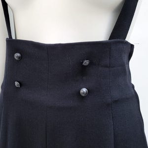 May include: A pair of high-waisted black trousers featuring two vertical rows of two small, round, dark buttons on the front waistband. The garment includes attached black suspender straps that extend upward from the high-rise waistline.