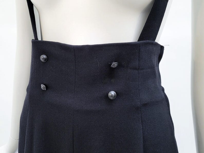May include: A pair of high-waisted black trousers featuring two vertical rows of two small, round, dark buttons on the front waistband. The garment includes attached black suspender straps that extend upward from the high-rise waistline.