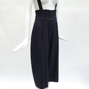 May include: High-waisted black trousers featuring wide shoulder straps that function as suspenders. The garment has a structured waistband with four small black buttons arranged in two vertical pairs on the front and a cropped leg length.