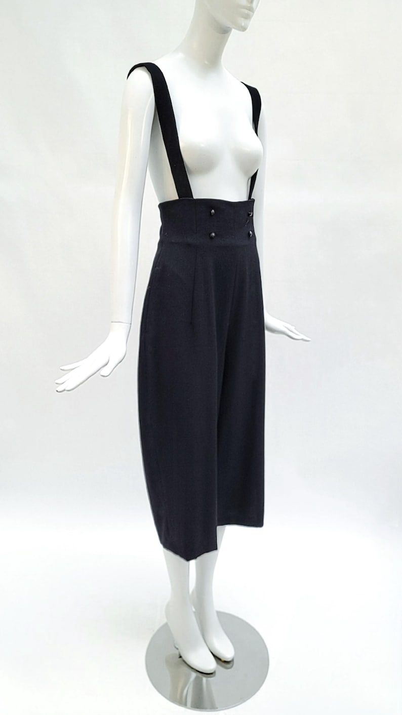 May include: High-waisted black trousers featuring wide shoulder straps that function as suspenders. The garment has a structured waistband with four small black buttons arranged in two vertical pairs on the front and a cropped leg length.
