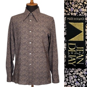 May include: A long-sleeved button-down shirt with a black and white floral pattern. The shirt has a pointed collar and a single button closure. The label reads "Jean Devy Made in France".