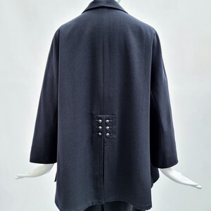 May include: A dark charcoal gray jacket featuring a structured collar and long sleeves. The back of the garment displays a central vertical seam with a decorative panel containing six small, round, metallic silver buttons arranged in two columns.