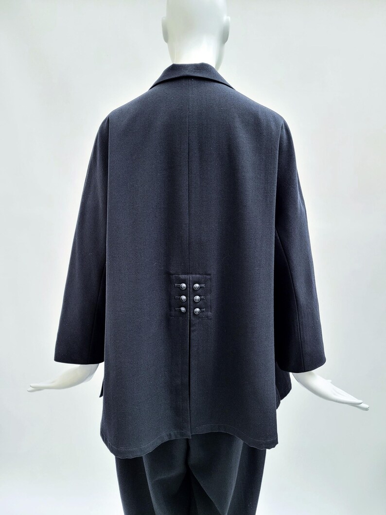May include: A dark charcoal gray jacket featuring a structured collar and long sleeves. The back of the garment displays a central vertical seam with a decorative panel containing six small, round, metallic silver buttons arranged in two columns.