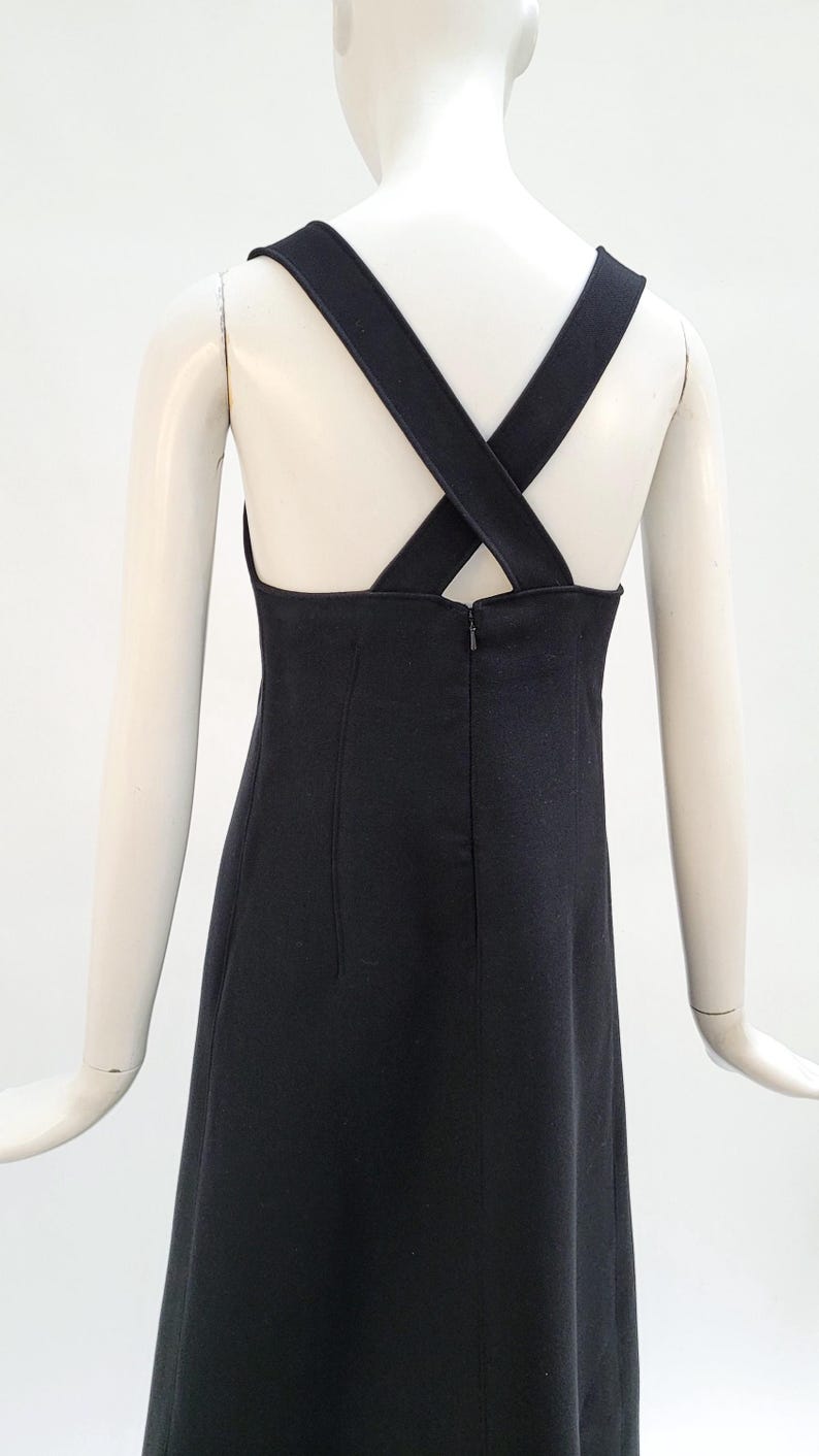 Pu&ograve; includere: Abito nero dal design semplice, con spalline incrociate sulla schiena. L'abito ha un corpetto aderente e una gonna dritta, con chiusura a zip sul retro. L'abito &egrave; esposto su un manichino.