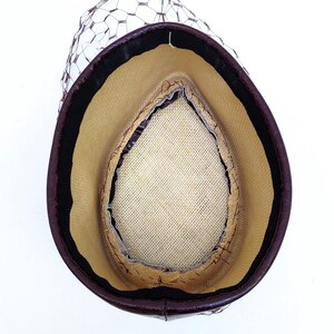May include: A teardrop-shaped hat viewed from the interior, featuring a dark brown outer rim, a beige woven straw-like lining, a black inner sweatband, and a dark brown netting veil attached to the exterior edge of the hat structure.