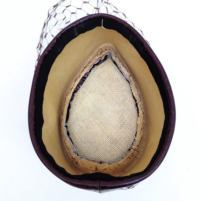 May include: A teardrop-shaped hat viewed from the interior, featuring a dark brown outer rim, a beige woven straw-like lining, a black inner sweatband, and a dark brown netting veil attached to the exterior edge of the hat structure.