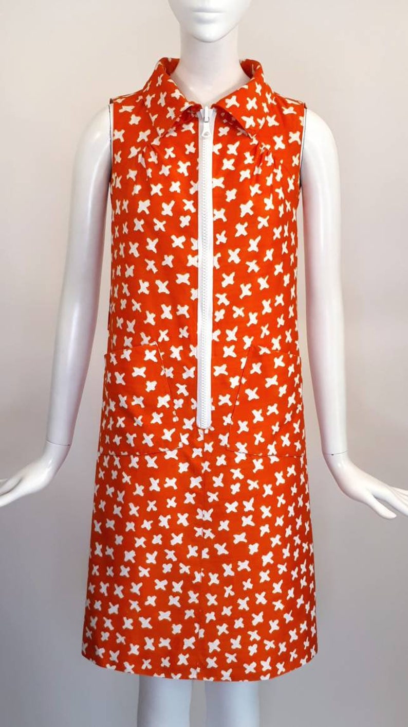 May include: A sleeveless orange dress with a white zipper and white geometric pattern. The dress has a white collar and two pockets.