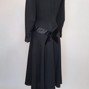 Puede incluir: Un vestido negro con un corpi&ntilde;o ajustado y una falda acampanada. El vestido tiene una faja de sat&eacute;n negro que se envuelve alrededor de la cintura y se ata en un lazo en la espalda. El vestido est&aacute; en un maniqu&iacute; de madera.