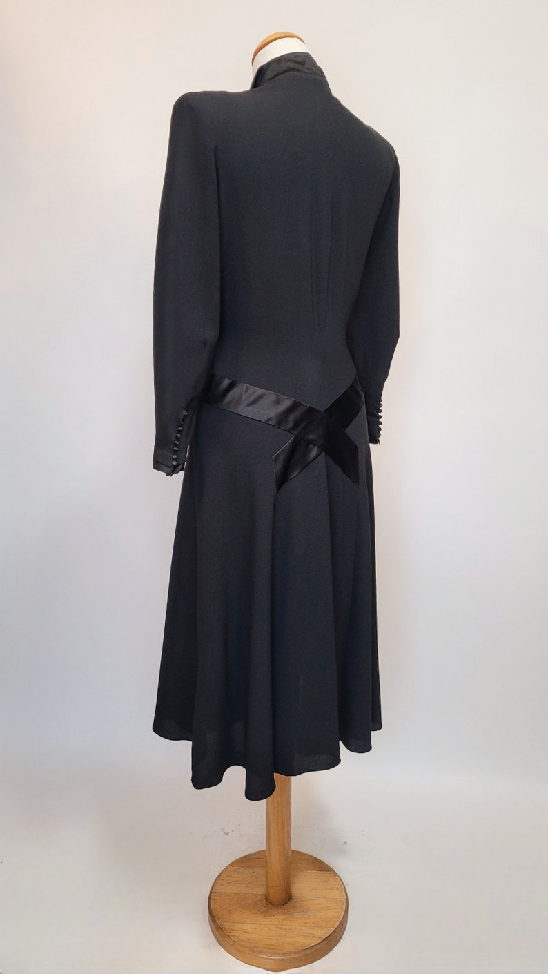 Puede incluir: Un vestido negro con un corpi&ntilde;o ajustado y una falda acampanada. El vestido tiene una faja de sat&eacute;n negro que se envuelve alrededor de la cintura y se ata en un lazo en la espalda. El vestido est&aacute; en un maniqu&iacute; de madera.