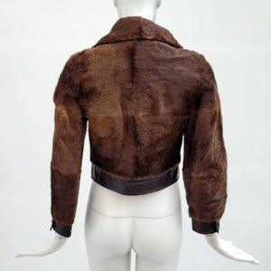 May include: A cropped brown fur jacket with a dark brown leather band at the waist and cuffs. The jacket has a collar and long sleeves. The back of the jacket is visible, showcasing the texture of the fur.