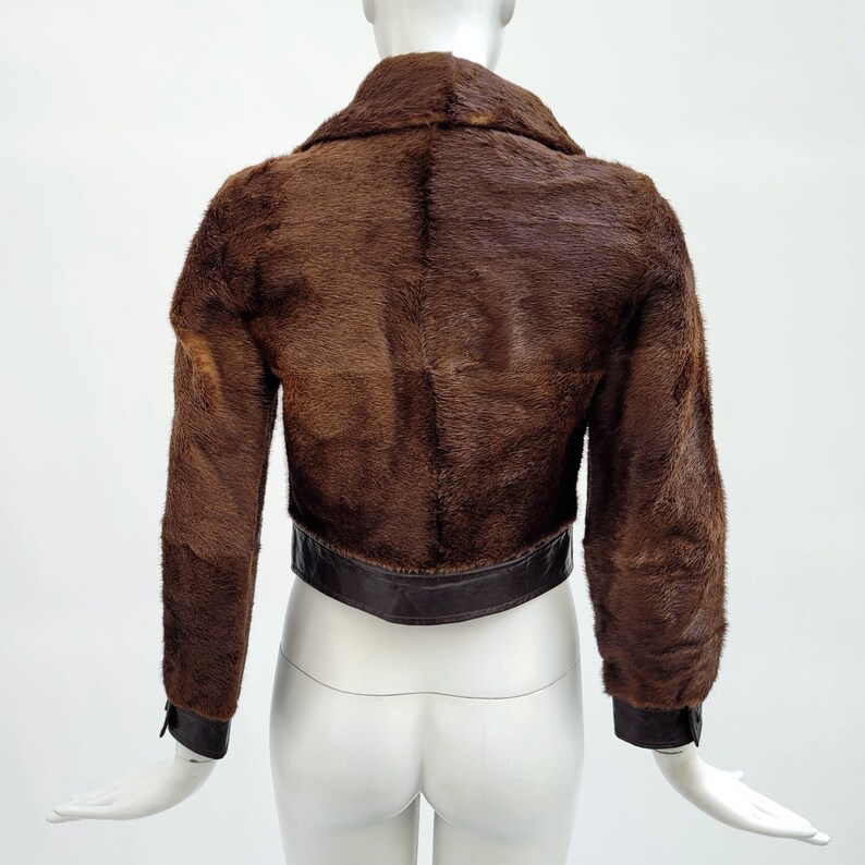 May include: A cropped brown fur jacket with a dark brown leather band at the waist and cuffs. The jacket has a collar and long sleeves. The back of the jacket is visible, showcasing the texture of the fur.