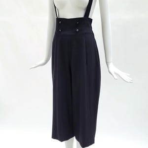 May include: A pair of dark navy blue high-waisted trousers with integrated suspender straps. The trousers feature a wide-leg cropped silhouette, a structured waistband with four small circular buttons arranged in two rows, and vertical pleats.