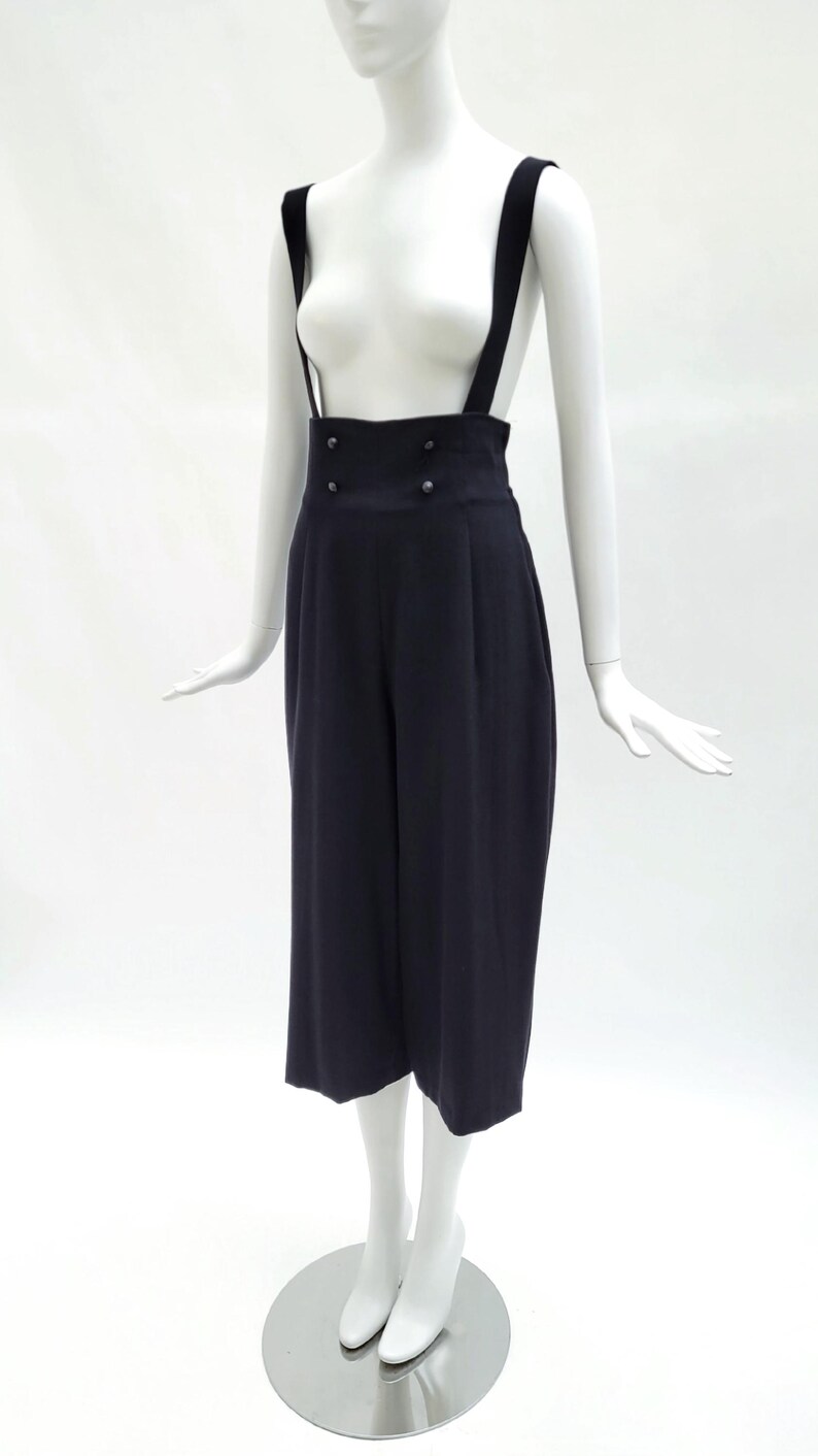 May include: A pair of dark navy blue high-waisted trousers with integrated suspender straps. The trousers feature a wide-leg cropped silhouette, a structured waistband with four small circular buttons arranged in two rows, and vertical pleats.