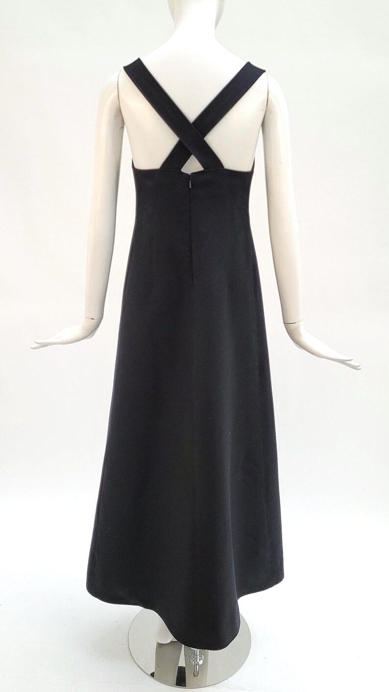 Pu&ograve; includere: Un abito nero lungo fino al pavimento, con scollatura all'americana e spalline incrociate sulla schiena. L'abito &egrave; esposto su un manichino su sfondo bianco. L'abito ha un design semplice ed elegante.