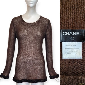May include: Brown knitted sweater with a loose fit and a delicate open weave. The sweater has a round neckline and long sleeves. The bottom hem and cuffs are trimmed with a beaded detail. The sweater has a label that reads "CHANEL" and "MADE IN ITALY".