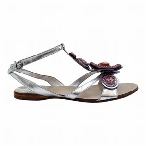 Peut inclure: Sandales en cuir argenté avec une fleur décorative. Les sandales présentent un design en T et une fermeture à boucle. La fleur est ornée de pierres roses et violettes. La semelle est en cuir marron.