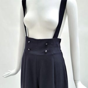 May include: High-waisted black trousers featuring wide shoulder straps and a double-breasted front closure with four small, round, dark buttons. The garment has a structured waistband and vertical pleats extending down the front of the legs.