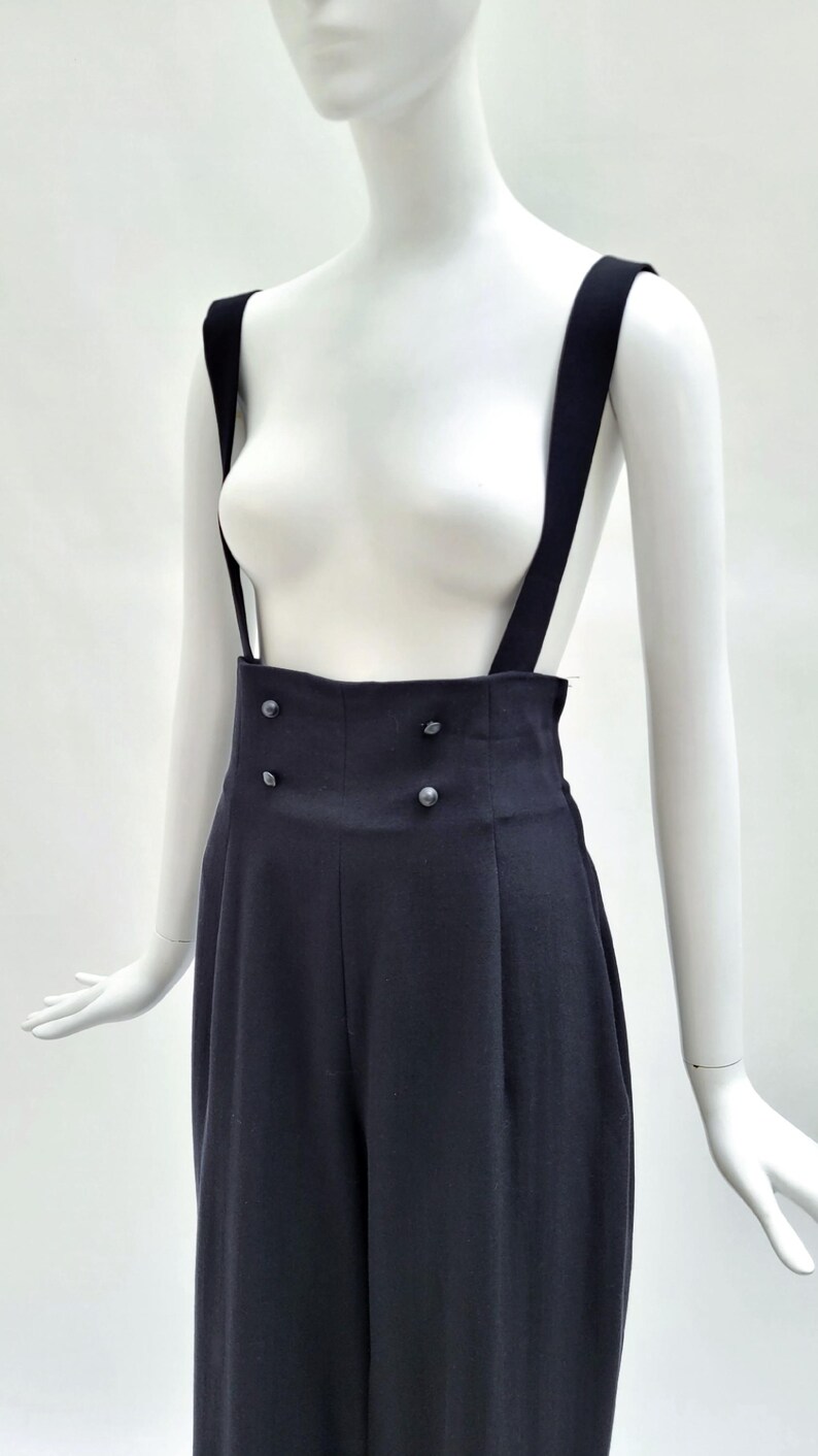 May include: High-waisted black trousers featuring wide shoulder straps and a double-breasted front closure with four small, round, dark buttons. The garment has a structured waistband and vertical pleats extending down the front of the legs.