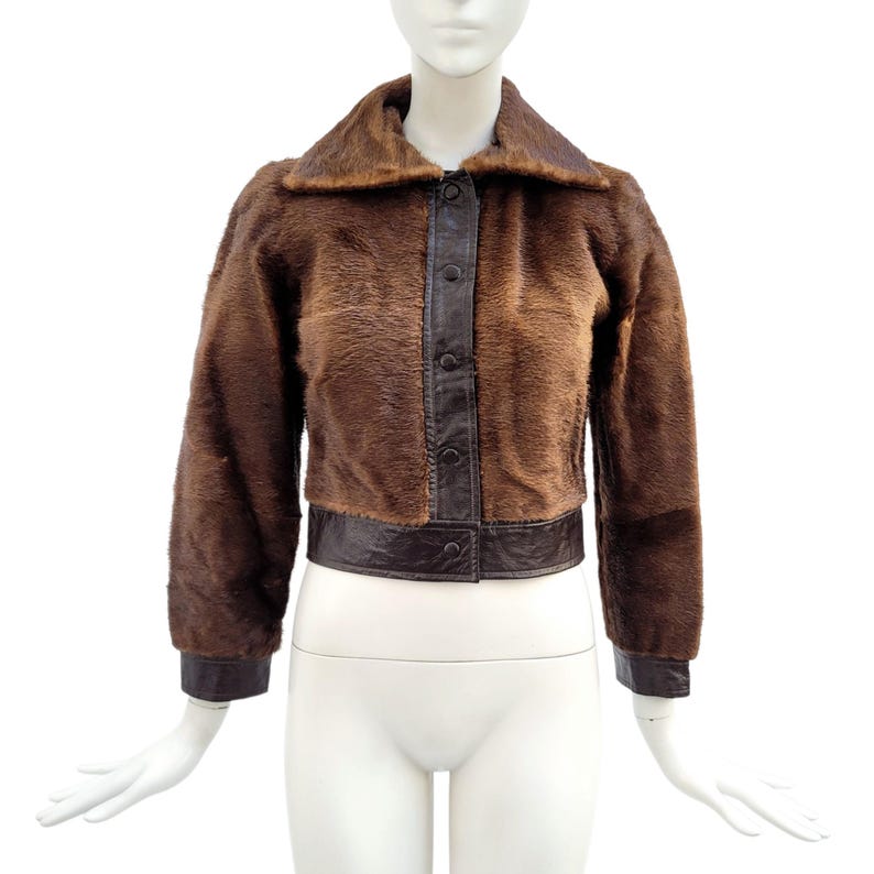 May include: A brown fur jacket with a dark brown leather collar, trim, and button closure. The jacket has long sleeves and is displayed on a mannequin. The jacket is a vintage style.