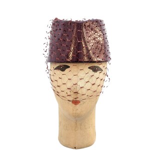 May include: A structured pillbox hat in a deep metallic burgundy with a textured, reptilian-like finish. The hat is adorned with a fine, matching burgundy netting veil that drapes over the front, creating a delicate, layered appearance.