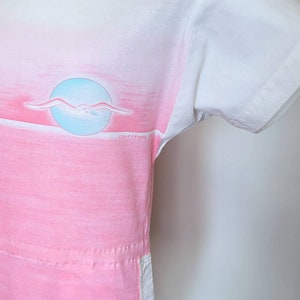 May include: A white short-sleeved t-shirt with a pink and blue ombre design. The shirt features a seagull flying over a blue circle and the text "courr&egrave;ges paris" in the bottom right corner.