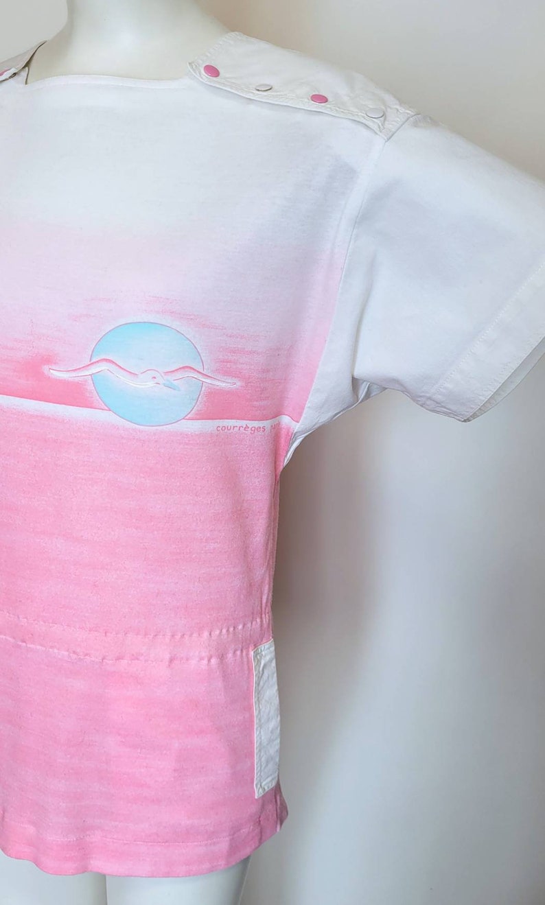 May include: A white short-sleeved t-shirt with a pink and blue ombre design. The shirt features a seagull flying over a blue circle and the text "courr&egrave;ges paris" in the bottom right corner.