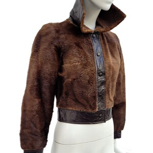 May include: A brown fur jacket with a high collar and leather detailing. The cropped jacket has a leather band at the waist and cuffs. The front features a leather panel with buttons. The jacket is displayed on a mannequin.
