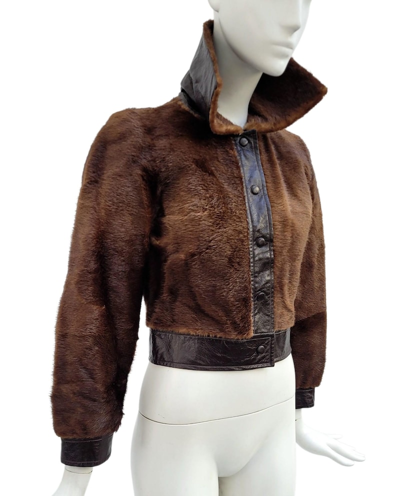 May include: A brown fur jacket with a high collar and leather detailing. The cropped jacket has a leather band at the waist and cuffs. The front features a leather panel with buttons. The jacket is displayed on a mannequin.