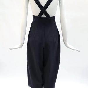 May include: A pair of high-waisted black trousers featuring wide legs that fall to a cropped length. The garment includes thick, integrated shoulder straps that cross over the back, creating a distinct X-shape design against the upper torso.