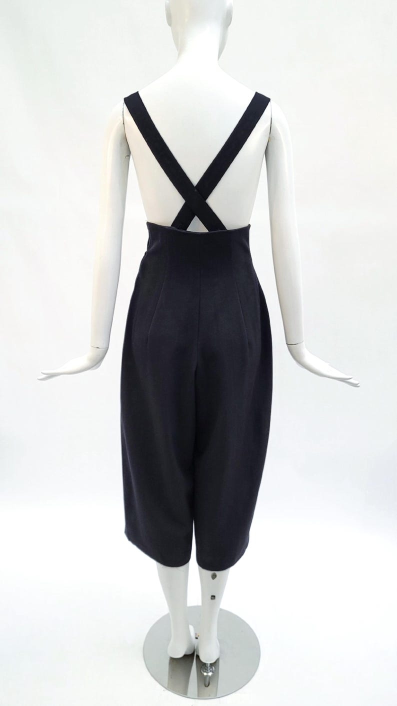 May include: A pair of high-waisted black trousers featuring wide legs that fall to a cropped length. The garment includes thick, integrated shoulder straps that cross over the back, creating a distinct X-shape design against the upper torso.