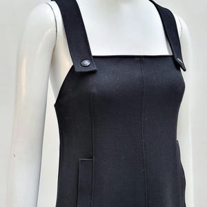 Pu&ograve; includere: Un abito nero con spalline larghe e scollatura quadrata. L'abito presenta due tasche frontali e un dettaglio a bottone sulle spalline. Il tessuto sembra essere un materiale di peso medio.